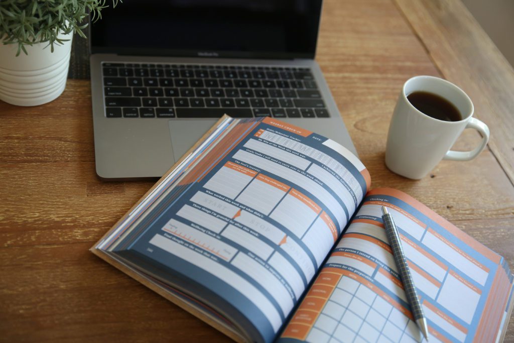 An image of a Story of My Life Journal, which can be used for time blocking, on a wooden desk, with a black coffee, opened laptop, and indoor plant nearby.